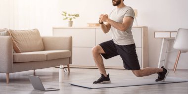 Atlet evde antrenman yapıyor, covid-19 sırasında yeni normal ve çevrimiçi dersler. Neşeli Arap sakallı genç adam oturma odasının içinde bacaklar için hamleler yapıyor, yerde dizüstü bilgisayara bakıyor, su şişesinin yanında.