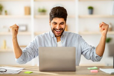 Aşırı sevinçli Arap girişimci iş başarısını kollarını kaldırarak kutluyor, dizüstü bilgisayar ekranına heyecanla bakıyor. Yumruklarını sıkan genç bir adam, evde oturuyor.