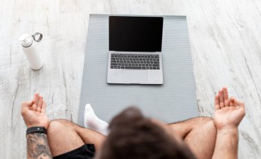 Kafkasyalı genç adam Lotus pozisyonunda meditasyon yapıyor yerde minder üzerinde oturuyor, boş ekranla su şişesi ve üst manzara ile PD 'ye bakıyor. Spor blogu, yoga sınıfı ve koçla egzersiz, covid-19 salgını.