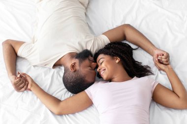 Smiling happy loving young african american lady and guy enjoy romantic moment lie on comfortable bed in bedroom, top view, empty space. Love, family, weekend together, couple enjoy time together