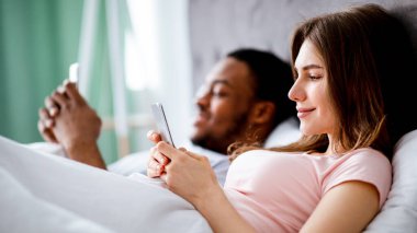 Gadget bağımlılığı konsepti. Genç, çok ırklı bir aile yatakta yatarken akıllı telefon kullanıyor. Kafkasyalı eş ve siyah kocası internette sörf yapıyor ya da oyunlar oynuyor, sosyal medya paylaşımları okuyor, yer kopyalıyor.