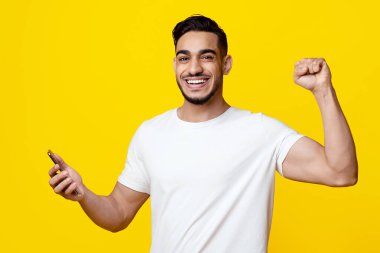 Beyaz tişörtlü, mutlu, mutlu, cep telefonu tutan, elini kaldıran, gülümseyen, sarı stüdyo arka planı, kopyalama alanı.