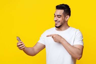 Happy bearded millennial arabic guy pointing at mobile phone screen and smiling, having video chat with friends, using mobile app for telecommunication, yellow studio background, copy space