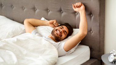 Dinlenmiş Arap adam uyanıyor ve ellerini uzatıyor, iyi uyuyan adam yatakta uzanıyor, rahat yatak odasında uyanıyor ve gözleri kapalı gülümsüyor. İyi uykular, tatlı rüyalar ve eğlence konsepti.