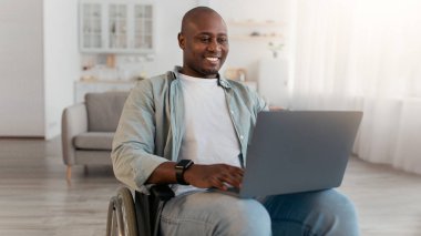 Modern iletişim, online çalışma, internette sörf ve sosyal ağlarda sohbet. Tekerlekli sandalyede sakat kalmış, gülümseyen Afrikalı Amerikalı adam bilgisayara bakıyor, oturma odasında mesaj yazıyor.