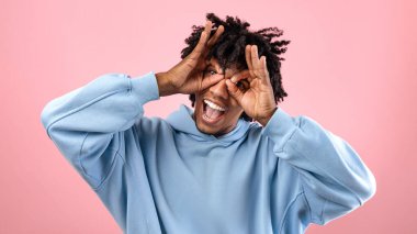 Aptal siyah genç adam parmaklarıyla uçak gözlüğü yapıyor, yüzünü buruşturuyor, pembe stüdyo arka planında komik bir ifade takınıyor. Neşeli Afrikalı Amerikalı ergen şakaları, kaygısız ve komik olmak