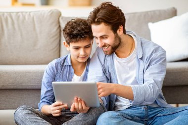 Baba ve oğul evde tablet bilgisayar kullanıyor, yeni başvuru formlarını kontrol ediyor, halının üzerinde oturuyor, oturma odasında kanepeye yaslanıyor, boş alan yaratıyor. Modern teknolojiler ve eğlence aktiviteleri konsepti