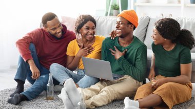 Mutlu siyahi arkadaşlar dizüstü bilgisayar kullanan genç erkekler ve kadınlar, oturma odasında oturmuş, video veya film seyrediyor ve gülüyorlar, Afrikalı Amerikalı çiftler evlerinde video konferansı yapıyorlar.