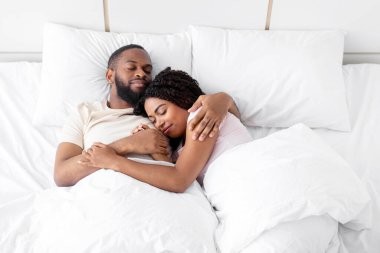 Loving happy peaceful millennial african american man hugs his woman and sleeps on comfortable bed together in bedroom, top view, empty space. Cozy home, rest and relaxation at weekend, health care