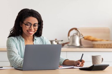 Güvenlik, yeni normallik, evde çalışma araçları, covid-19 salgını. Gülümseyen bin yıllık Afro-Amerikan gözlüklü çekici kadın notlar alıyor, modern mutfak içindeki bilgisayar ekranlarına bakıyor.