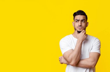 Düşünceli, yakışıklı, gündelik kıyafetli orta yaşlı bir adam çenesine dokunuyor ve sarı stüdyo arka planında reklam için fotokopi alanına bakıyor.