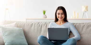 Yeni normal, sosyal ağlarda sörf, evde başlangıç ve iş, blog, uzaktan çalışma, covid-19 salgını. Minimalist oturma odasındaki kanepede dizüstü bilgisayarda yazan neşeli, bin yıllık Arap kadın.