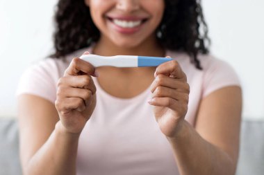 Harika haberler, olumlu duygular, aile planlaması, yumurtlama. Mutlu, heyecanlı, binlerce yıllık Afro-Amerikan kadınının portresi oturma odasının iç kısmında pozitif hamilelik testine bakıyor.