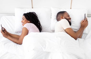 Cheerful glad young black lady and guy suffer from addiction to phone, social networks and online games. Couple surfing in internet, lies on bed in bedroom interior. Relationship problems, new normal