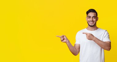 Cheerful middle eastern millennial guy in casual outfit pointing at copy space over yellow studio background, showing nice advertisement or recommending exciting deal, panorama