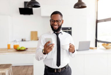 Smiling Black Business Man In White Shirt And Eyglasses Using Smartphone And Drinking Coffee While Standing At Home Office, Browsing Internet Or Social Networks, Enjoying Free Time And Break From Work