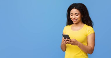 Havalı bir alet. Heyecanlı Afrikalı Amerikalı kadın akıllı telefon kullanıyor, mesajlaşıyor, internette geziniyor ya da sosyal ağlarda geziniyor. Mavi stüdyo arka planında fotokopi çekiyor.