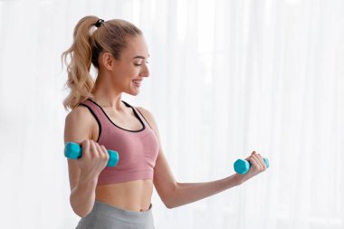 Neşeli Avrupalı genç sarışın kadın evde dambılları kaldırıyor, tek başına çalışıyor, salgın sırasında cesetle ilgileniyor. El egzersizleri ekipman ve sağlıkla yapılır.