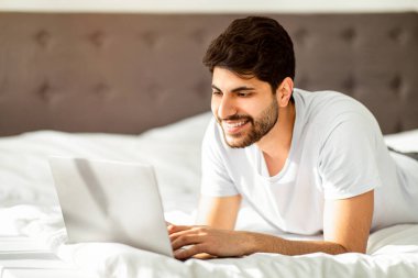 Randevu sitesi, cihazla boş zaman geçirmek, internette sörf yapmak ve evde çalışmak. Beyaz tişörtlü mutlu Arap adam yatak odasında iç tarafta uzanıyor ve dizüstü bilgisayarda yazıyor.
