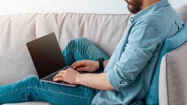 Rahat Arap adam boş ekranlı dizüstü bilgisayar kullanıyor, oturma odasında koltukta dinleniyor, iç mekan, boş yer, maket. Boş ekran ile bilgisayara bakan genç adam