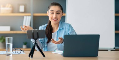 Video konferansı, evde bilgisayar öğretmeniyle e-öğrenme. Evde eğitim, uzaktan eğitim, online eğitim. Kara tahtanın yanındaki arkadaş canlısı Arap kadın akıllı telefon webcam, laptop 'a bakıyor.