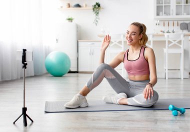 Modern fitness blogcusu online sınıf kumandasıyla egzersiz videosu kaydediyor. Mutlu sarışın kadın mindere oturur, oturma odasının içinde el sallar halterler ve bir şişe su ile el sallar.