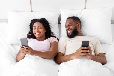 Glad smiling young black female and male, lying on bed, typing on phones, chatting, playing online game in bedroom interior. Great news, sale, offer, app and device addiction, new normal, top view