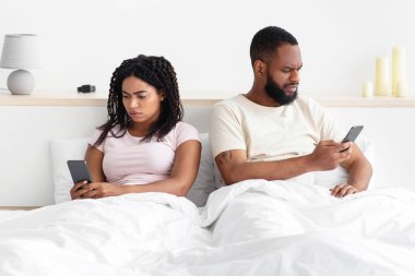 Offended sad young african american guy ignores lady and typing on smartphones on comfortable bed in bedroom. Relationship problems, quarrel, new app. Surfing couple searching, chatting in internet
