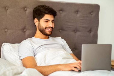 Uzaktan kumanda. Dizüstü bilgisayarda çalışan mutlu Arap serbest çalışanı evde yatakta otururken internette geziniyor. Sabahları bilgisayar kullanan bir adam. Serbest kariyer ve yaşam tarzı konsepti