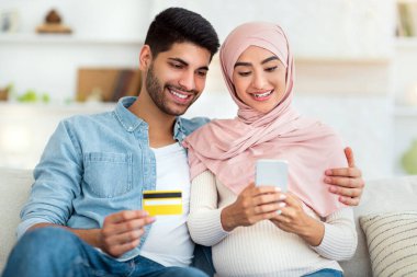 Mutlu Müslüman çift satın almak için mobil uygulama kullanıyor, kredi kartıyla bebek için bir şeyler alıyor, ev içi koltukta oturuyor, yer kopyalıyor.