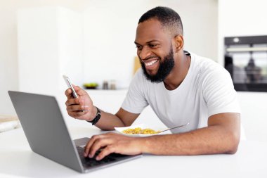 Gülümseyen siyah adamın portresi akıllı telefon ve bilgisayar kullanarak mutfakta kahvaltı etmesi, neşeli Afrikalı Amerikalı erkek masada oturuyor, klavyede yazıyor ve yemek yiyor, bedava fotokopi çekiyor.