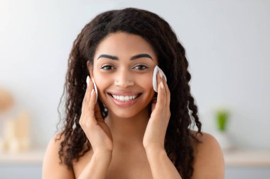 Makyaj malzemelerinin temizlenmesi, cilt bakımı ve evdeki günlük prosedürler. Mutlu bin yıllık Afro-Amerikan kadın beyaz havluyla kozmetik ürünlerini gri duvar arkasında pamuk yastıkla kaldırın, kapatın.