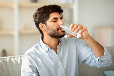 Genç Arap adam camdan tatlı su içiyor, oturma odasındaki kanepede oturuyor. Sağlık, diyet, evde düzgün beslenme, sabah ve ev yaşamında susuzluk, hidrasyon konsepti