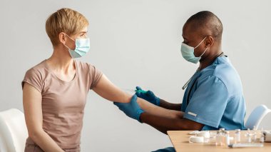 Tıbbi profesyonel bakım, sertifika için koronavirüse karşı aşı. Orta yaşlı, koruyucu maskeli Afrikalı Amerikalı doktor masadaki yetişkin kadına ilaç ve ilaç veriyor.