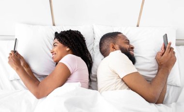 Glad young african american guy and lady chatting on smartphones lie on comfortable bed in bedroom. Game, social networks and devices addiction. Problems at home, hiding, cheating during covid-19