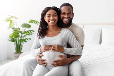 Satisfied smiling handsome millennial african american guy hugs pregnant lady with belly, sit on bed in white bedroom interior. Planning child, future family, waiting for baby in cozy home, free space