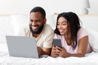 Cheerful young african american husband and wife with smartphone, lie on comfortable bed and look at laptop in bedroom interior. Video call, communication remotely, chat during COVID-19 lockdown