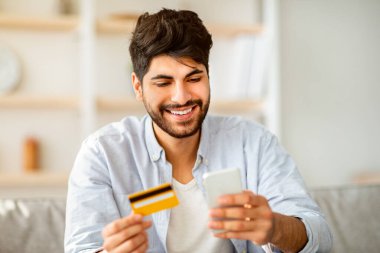 Çevrimiçi alışveriş. Mutlu Arap adam internetten ürün sipariş etmek için telefon ve kredi kartı kullanıyor, evdeki kanepede oturuyor. Ödeme doğrulama ve mobil bankacılık kavramı