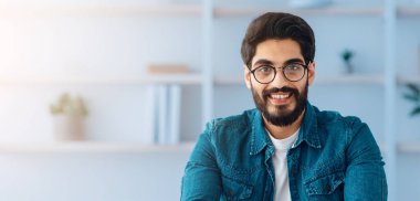 Mutlu Toung Arap kendini beğenmiş girişimci iç ofis masasında oturuyor ve kameraya gülümsüyor. İş yerinde poz veren yakışıklı iş adamı, kopyalama alanı