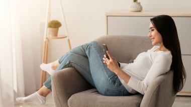 Evde dinlenmek, sosyal ağlar, modern sohbetler, internette sörf yapmak, boş zamanların tadını çıkarmak. Gülümseyen güzel Arap kadın minimalist oturma odasında, koltukta akıllı telefondan mesaj okuyor.