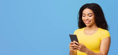 Mutlu Afro-Amerikalı kadın akıllı telefon mesajları ve iletişim ağlarını kullanıyor mavi stüdyo arka planında duruyor, boş alan. Telefondaki mobil uygulamayı kullanan siyah genç bayan.