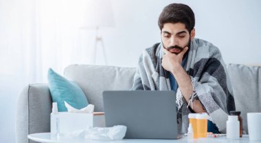 Hasta Arap erkek çalışan virüsten muzdarip ama işe ihtiyacı var, dizüstü bilgisayarda çalışıyor ve grip için hap alıyor. Evdeki kanepede oturan ve bilgisayar, kopya alanı kullanan adam.