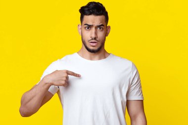 Confused handsome bearded young muslim guy in white t-shirt pointing at himself, having puzzled face expression, posing alone on yellow studio background, copy space