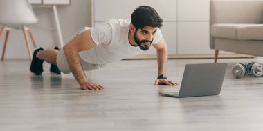 Karantinada sağlık ve vücut bakımı. Evde antrenman. Minimalist yatak odasında şınav çeken çekici 30 'lu yaşlarda genç bir Arap, yerde dizüstü bilgisayara bakıyor, dambıl ve su şişesinin yanında.