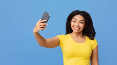 Akıllı telefondan özçekim yapan pozitif siyahi kadın mavi stüdyo arka planında poz veriyor ve eğleniyor. Kendi portreleri konsepti için sosyal medya, blog, mobil uygulama