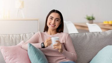 En sevdiğiniz içecekle dinlenin, tembellik, kahve molası, dinlenin, hafta sonları evde boş zaman geçirin, Cvid-19 tecrit. Gülen milenyum Müslüman kadın elinde bir fincan çayla minimalist oturma odasındaki kanepede oturuyor.