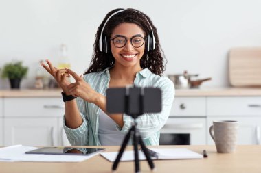 Sosyal mesafe, öğretmen ve öğretmenin uzaktan çalışması, modern blog ve çalışma. Gözlüklü, kulaklıklı, gülümseyen Afro-Amerikan kadın jestler, konuşmalar ve mutfakta tripodlarda web kameralarına bakıyor.