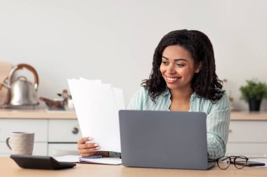 Ev muhasebesi, vergiler, yeni normal ve uzak bir iş, evde iyi eğlenceler. Beyaz minimalist mutfak iç mekânında, bilgisayar ve hesap makinesiyle çalışan mutlu, Afro-Amerikalı genç bir kadın.