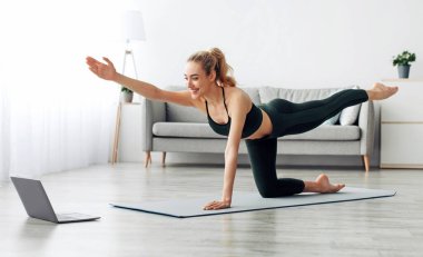 Modern spor, yeni normal, güvenli, aktif yaşam tarzı, çevrimiçi cihaz eğitimi. Spor giyimli neşeli genç bayan yoga yapıyor, tahta yapıyor, odanın içinde dizüstü bilgisayarla statik egzersiz yapıyor.