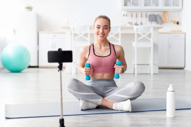 Neşeli Avrupalı bin yıllık sarışın fitness blogcusu webcam 'e bakar ve evde sosyal ağlar için halterli antreman videosu çeker. Modern blog, spor ve aygıt, yeni normal ve güvenli